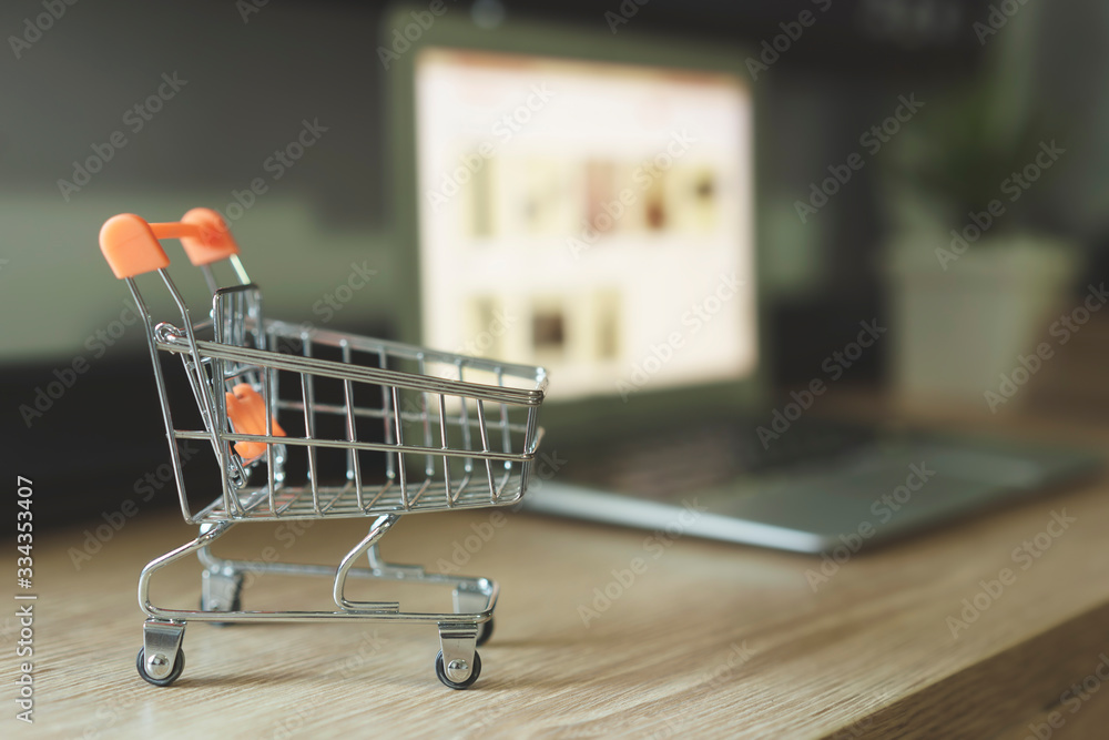 Online shopping concept, Shopping cart with blurry laptop on the desk Online shopping concept, Shopping cart with blurry laptop on the desk