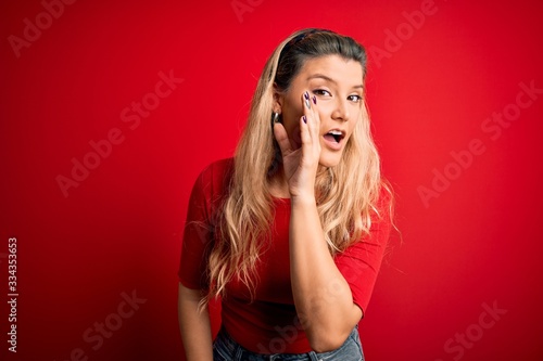 Young beautiful blonde woman wearing casual t-shirt standing over isolated red background hand on mouth telling secret rumor, whispering malicious talk conversation