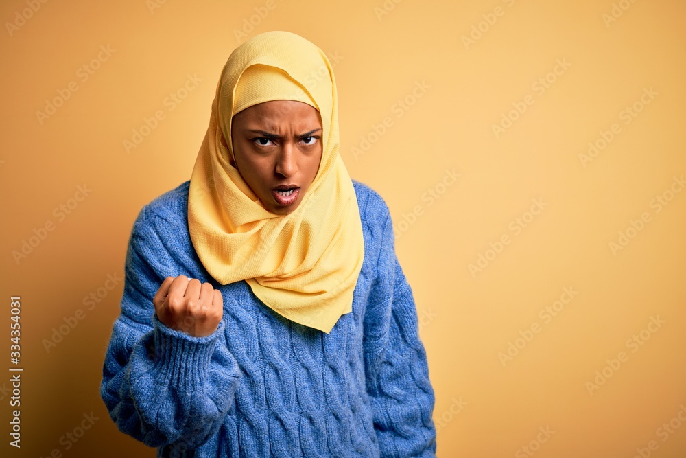 Young African American afro woman wearing muslim hijab over isolated yellow background angry and mad raising fist frustrated and furious while shouting with anger. Rage and aggressive concept. Young African American afro woman wearing muslim hijab over isolated yellow background angry and mad raising fist frustrated and furious while shouting with anger. Rage and aggressive concept.