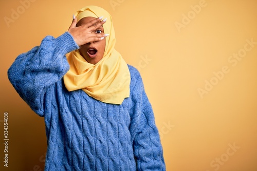 Young African American afro woman wearing muslim hijab over isolated yellow background peeking in shock covering face and eyes with hand, looking through fingers with embarrassed expression. © Krakenimages.com