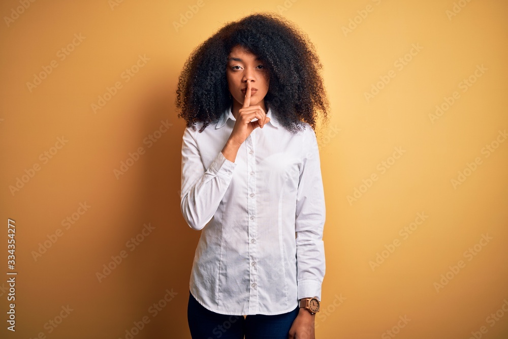 Young beautiful african american elegant woman with afro hair standing over yellow background asking to be quiet with finger on lips. Silence and secret concept. Young beautiful african american elegant woman with afro hair standing over yellow background asking to be quiet with finger on lips. Silence and secret concept.