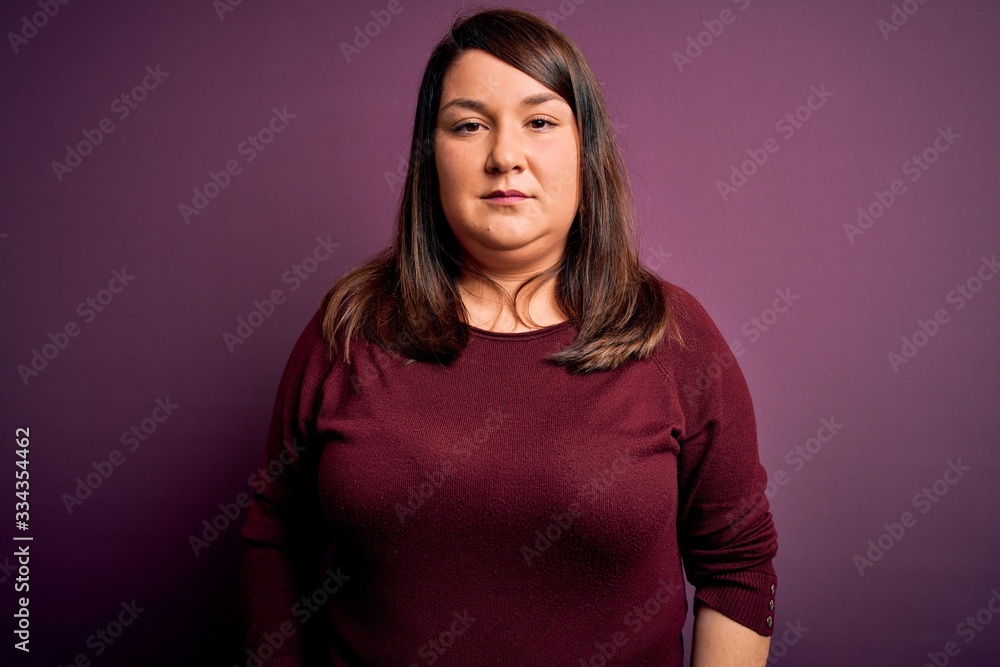 Obraz premium Beautiful brunette plus size woman wearing casual sweater over isolated purple background with serious expression on face. Simple and natural looking at the camera.
