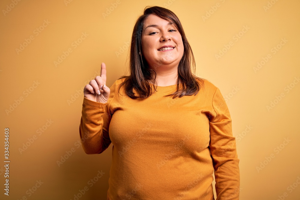 Beautiful brunette plus size woman wearing casual sweater over isolated yellow background showing and pointing up with finger number one while smiling confident and happy. Beautiful brunette plus size woman wearing casual sweater over isolated yellow background showing and pointing up with finger number one while smiling confident and happy.
