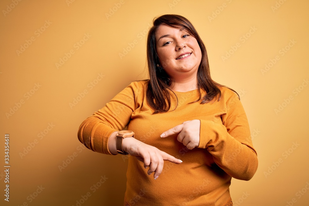 Obraz premium Beautiful brunette plus size woman wearing casual sweater over isolated yellow background In hurry pointing to watch time, impatience, upset and angry for deadline delay