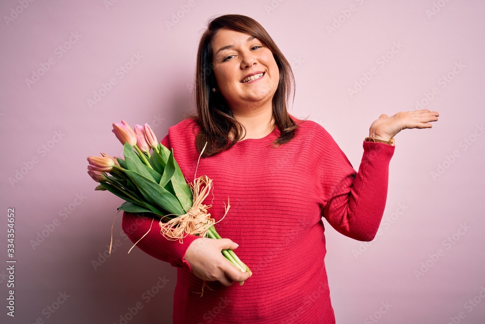 Beautiful plus size woman holding romantic bouquet of natural tulips flowers over pink background smiling cheerful presenting and pointing with palm of hand looking at the camera. Beautiful plus size woman holding romantic bouquet of natural tulips flowers over pink background smiling cheerful presenting and pointing with palm of hand looking at the camera.