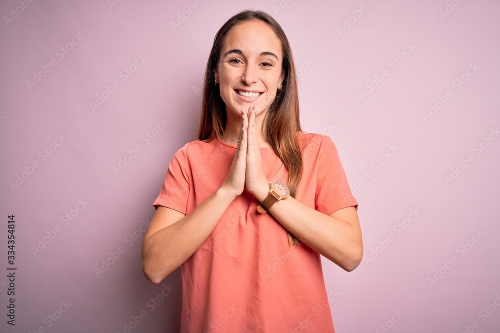 Young beautiful woman wearing casual t-shirt standing over isolated pink background praying with hands together asking for forgiveness smiling confident. Young beautiful woman wearing casual t-shirt standing over isolated pink background praying with hands together asking for forgiveness smiling confident.