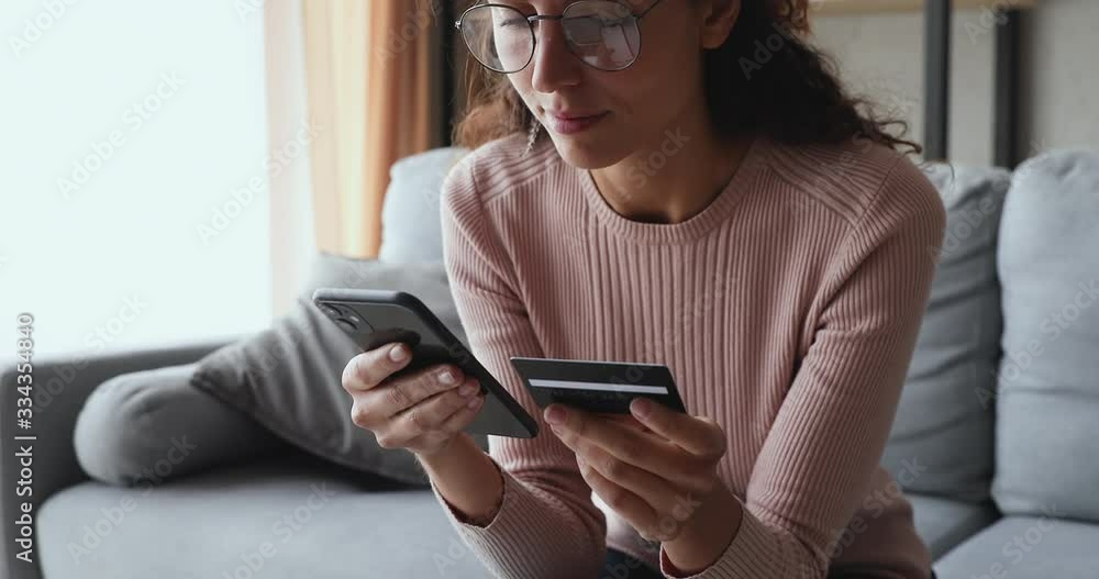 Smiling young woman customer holding credit card and smartphone sitting on couch at home. Happy female shopper using instant easy mobile payments making purchase in online store. E-banking app service