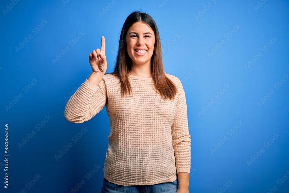 Young beautiful woman wearing casual sweater over blue background showing and pointing up with finger number one while smiling confident and happy.