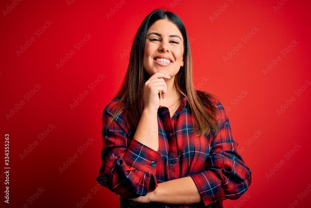 Young beautiful woman wearing casual shirt over red background looking confident at the camera with smile with crossed arms and hand raised on chin. Thinking positive. Young beautiful woman wearing casual shirt over red background looking confident at the camera with smile with crossed arms and hand raised on chin. Thinking positive.