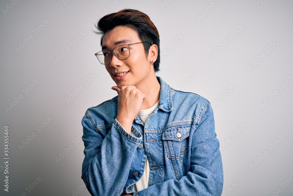 Young handsome chinese man wearing denim jacket and glasses over white background with hand on chin thinking about question, pensive expression. Smiling with thoughtful face. Doubt concept.
