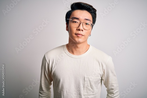 Young handsome chinese man wearing casual t-shirt and glasses over white background with serious expression on face. Simple and natural looking at the camera. © Krakenimages.com