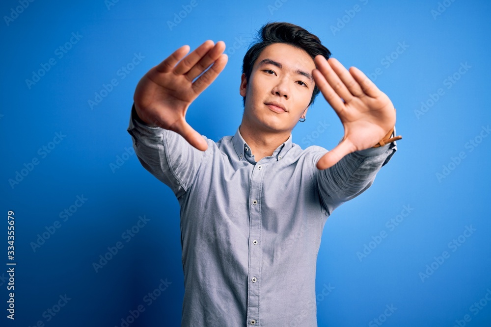 Naklejka premium Young handsome chinese man wearing casual shirt standing over isolated blue background doing frame using hands palms and fingers, camera perspective