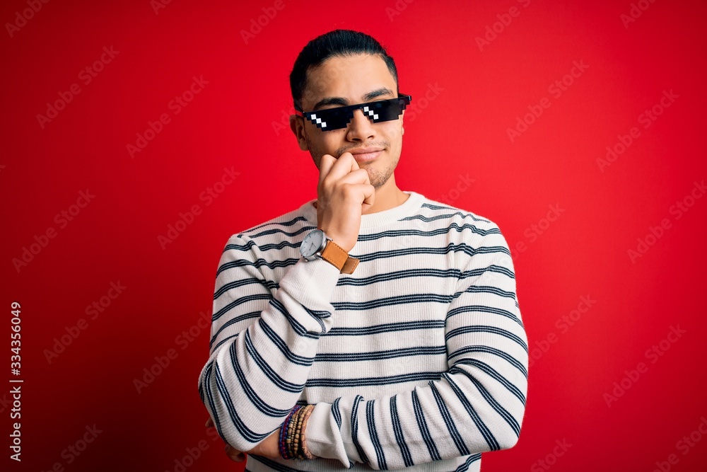 Obraz premium Young brazilian man wearing funny thug life sunglasses over isolated red background with hand on chin thinking about question, pensive expression. Smiling with thoughtful face. Doubt concept.
