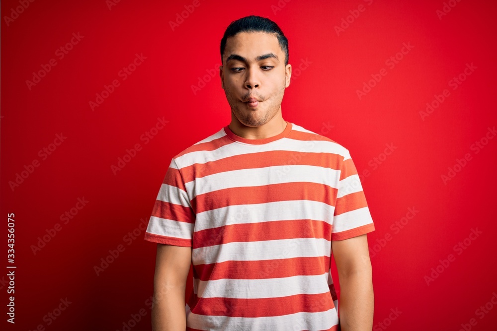 Young brazilian man wearing casual striped t-shirt standing over isolated red background making fish face with lips, crazy and comical gesture. Funny expression. Young brazilian man wearing casual striped t-shirt standing over isolated red background making fish face with lips, crazy and comical gesture. Funny expression.