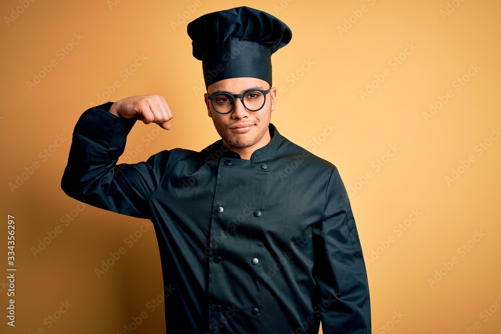 Young brazilian chef man wearing cooker uniform and hat over isolated yellow background Strong person showing arm muscle, confident and proud of power Young brazilian chef man wearing cooker uniform and hat over isolated yellow background Strong person showing arm muscle, confident and proud of power