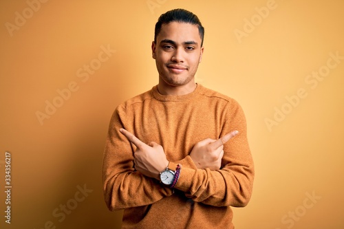 Young brazilian man wearing casual sweater standing over isolated yellow background Pointing to both sides with fingers  different direction disagree