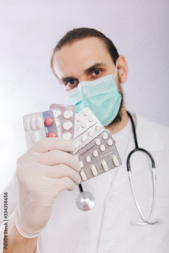 A doctor in a white coat, a medical mask and gloves holds pills. Types of drugs. Medical worker on a white background. Studio photo A doctor in a white coat, a medical mask and gloves holds pills. Types of drugs. Medical worker on a white background. Studio photo