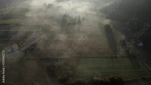 Aerial footage of hamlet, village Grahovo at sunrise through clouds and sun rays. Idyllic landscape. Small village in mountains, private houses, field, meadow, trees and beautiful shadows.