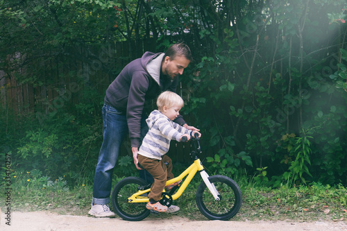 Cute blond boy learning to ride bicycle. Father teaching his little child to ride bike in spring summer park. Happy family moments. Time together dad and son. Candid lifestyle image.