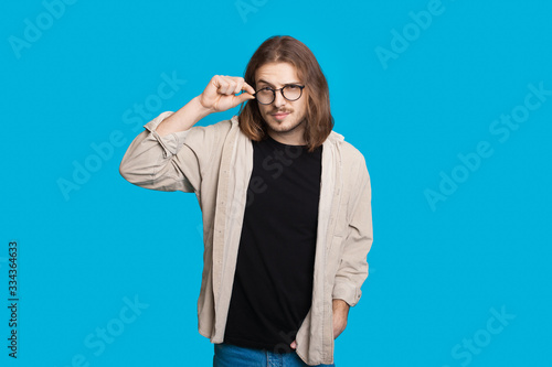 Caucasian man with long hair touching his eyeglasses and looking at camera on a blue wall