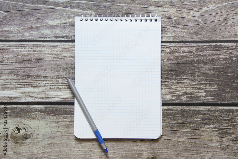 Notepad with a blank white sheet in a checker paper with ball pen lies on the background of wooden boards. Notepad with a blank white sheet in a checker paper with ball pen lies on the background of wooden boards.