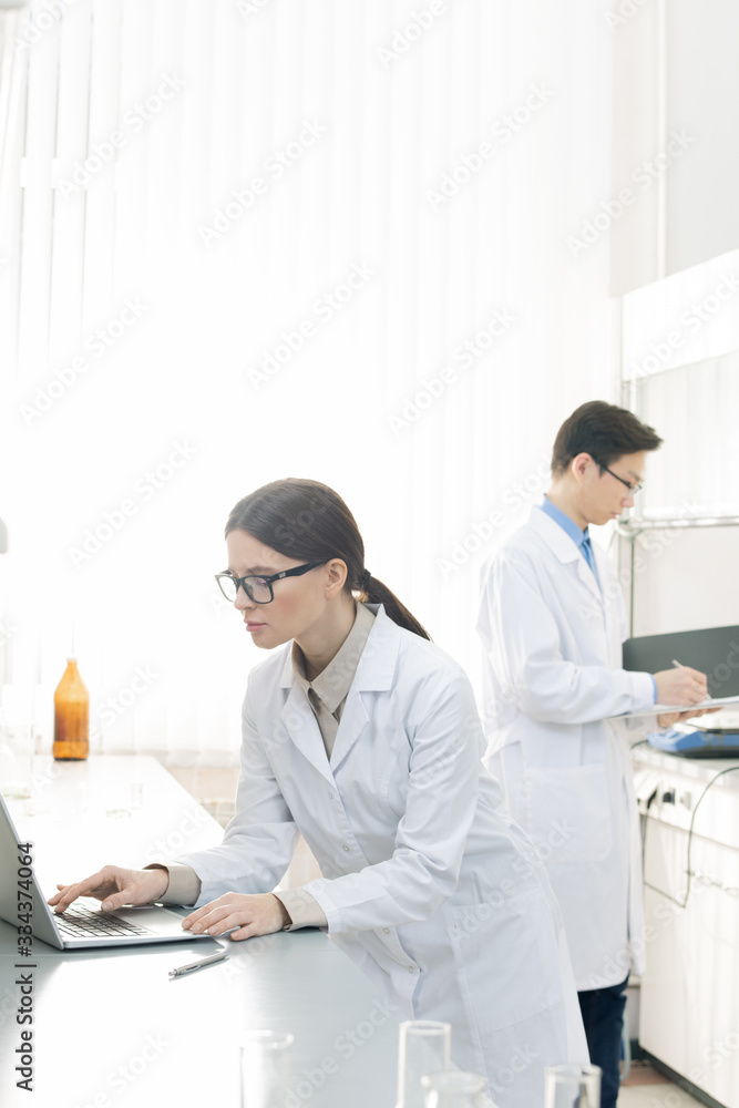 Pretty young lab worker in whitecoat and eyeglasses standing in front of laptop Pretty young lab worker in whitecoat and eyeglasses standing in front of laptop