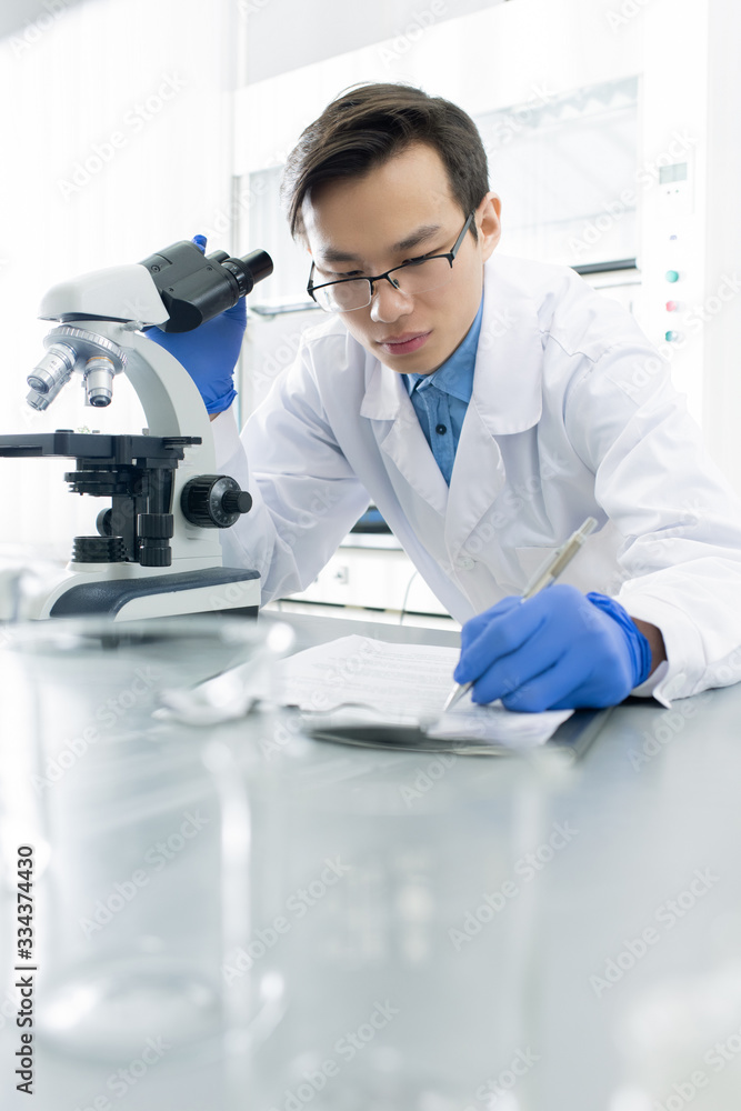 Chinese male lab worker in gloves and whitecoat making notes in notepad Chinese male lab worker in gloves and whitecoat making notes in notepad