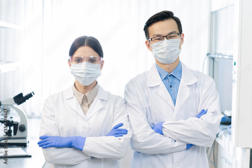 Two young masked intercultural scientists in whitecoats and gloves Two young masked intercultural scientists in whitecoats and gloves