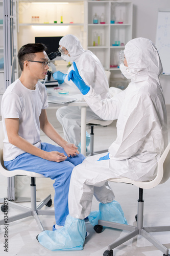 Young Chinese man sitting on chair in front of scientist in coveralls and gloves