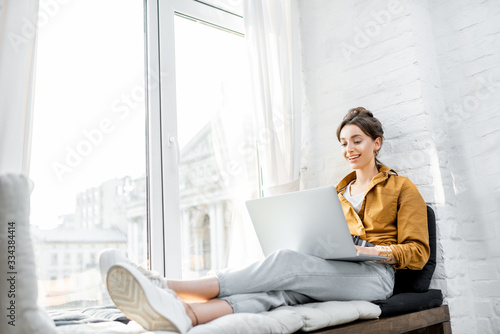 Young woman dressed casually working on laptop while sitting on the window sill at home. Work from home at cozy atmosphere concept photo