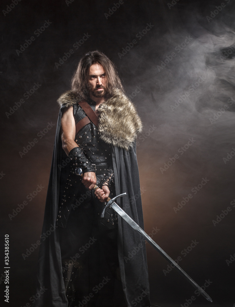 Man dressed in medieval armour and raincoat with longs word over smoke background. Courage fantasy warrior knight with long hair concept historical photo