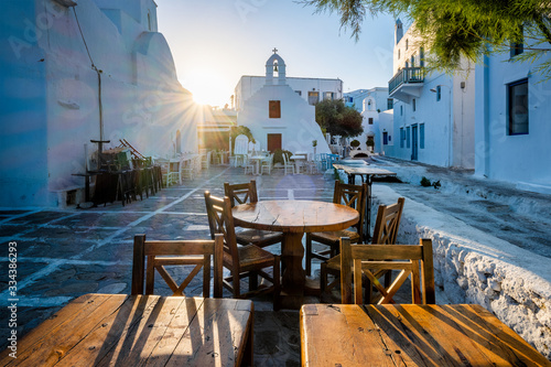 Fototapeta Naklejka Na Ścianę i Meble -  Tourist Greece scene - restaurant cafe table in picturesque scenic narrow streets with traditional whitewashed houses blue doors windows Chora town in famous tourist attraction Mykonos island, Greece