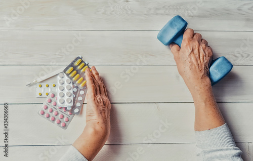 Senior hands with pills, drugs on table, dumb-bell. Woman wrinkled hands, thermometer, colorful tablets and dumb-bell, wooden background. Health care for the elderly, healthy living concept.