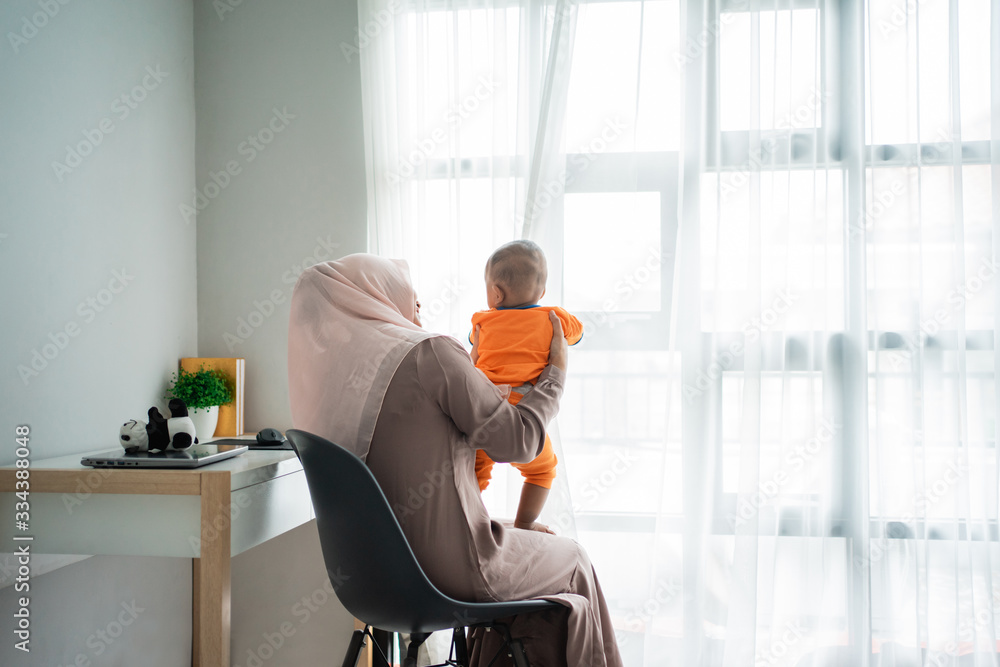 Asian moslem mother holding her little boy on lap when playing together front a window bedroom