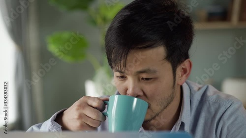 Work from Home. At close shot young Korean man with a mustache and a beard sits at a desk, quietly working on a laptop, drinks a hot drink from a cup in a cozy home environment.