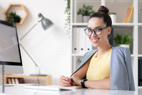 Happy young businesswoman in smart casual looking at you by desk