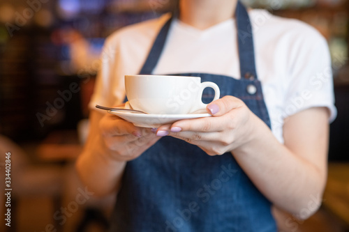 Young waitress in blue apron holding or carrying cup of tea or cappuccino