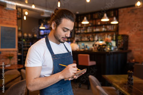 Young waiter in blue apron and white t-shirt writing down order of client