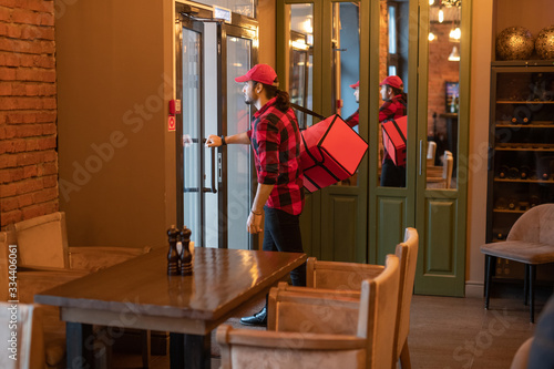 Young courier with big red bag on shoulder going to open the door of cafe