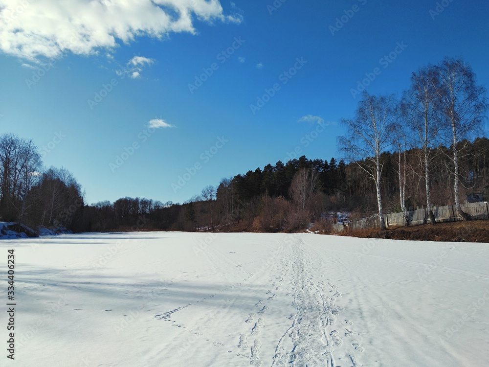 a spring Sunny landscape with a pond that has not yet thawed after winter hibernation through which there is a well trodden road against the background of beautiful trees and blue sky a spring Sunny landscape with a pond that has not yet thawed after winter hibernation through which there is a well trodden road against the background of beautiful trees and blue sky