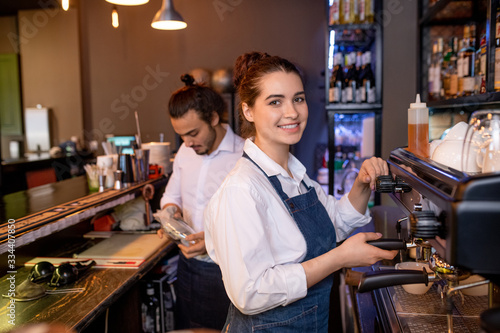 Happy young attractive waitress of cafe or restaurant preparing cappuccino