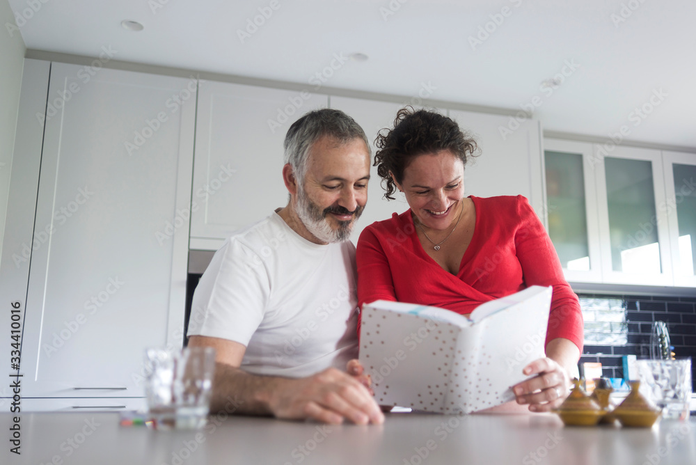 Obraz premium Happy pregnant woman with her husband sitting at home. Smiling couple consulting a book at the kitchen.