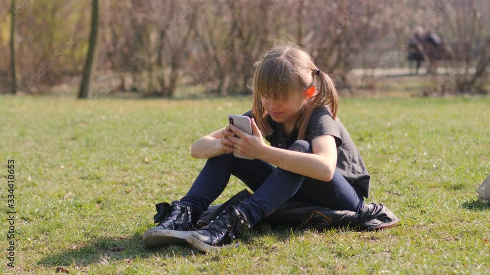 Lazy inactive kid girl with smartphone sit and lie on park lawn grass watching device diplay playing game application Lazy inactive kid girl with smartphone sit and lie on park lawn grass watching device diplay playing game application
