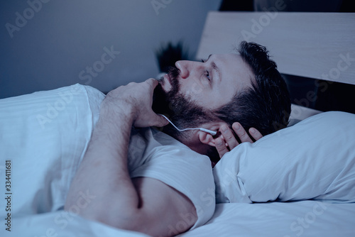 A lonely bearded dark-haired man lies on the bed at night and listens to music with a pensive expression on his face. Close-up, insomnia, bedroom, self-isolation.