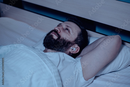 A lonely bearded dark-haired man lying on the bed at night and listening to music on wireless headphones with a dreamy expression on his face. Close-up, insomnia, bedroom, self-isolation.