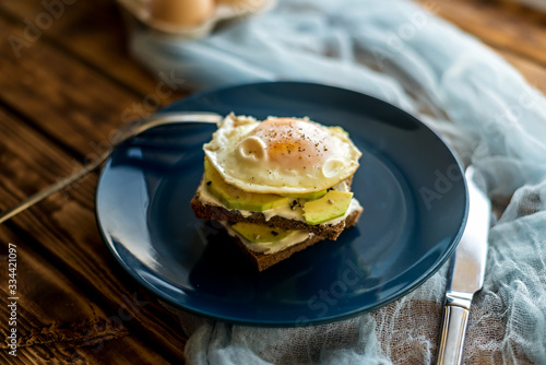 on a wooden table with blue cloth there is breakfast in a blue plate fried eggs on slices of dark bread with white sauce and avocado, in the background in the tray are raw eggs