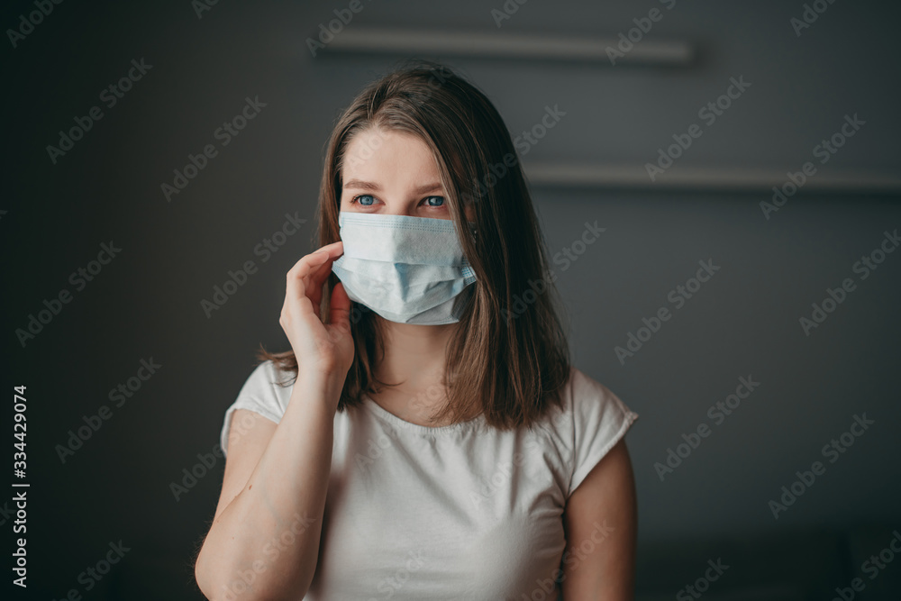 Young blonde white european blue-eyed woman at home on quarantine with medical mask on gray background. Coronavirus, illness, infection, quarantine, surgical bandage. Young blonde white european blue-eyed woman at home on quarantine with medical mask on gray background. Coronavirus, illness, infection, quarantine, surgical bandage.
