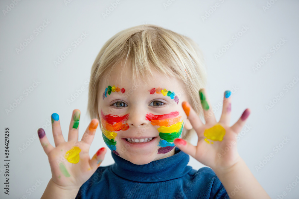 Beautiful blond toddler boy with rainbow painted on his face and messy hands Beautiful blond toddler boy with rainbow painted on his face and messy hands