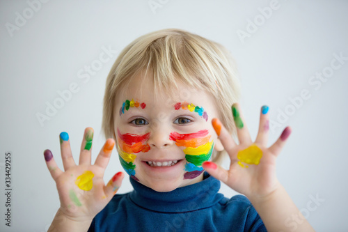 Beautiful blond toddler boy with rainbow painted on his face and messy hands