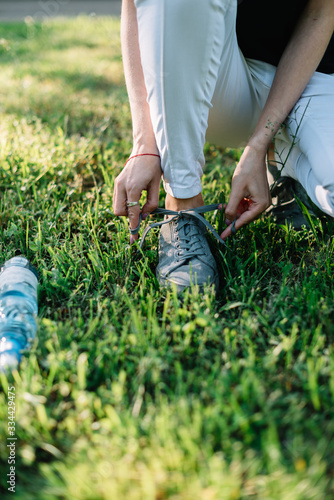 Girl athlete tying shoelaces on sneakers in a summer park. Nearby is a bottle of water